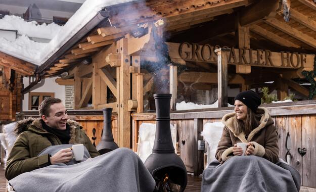Gäste genießen heiße Getränke im verschneiten Außenbereich des Boutique Hotel Salzburg vor einem Kamin, umgeben von traditioneller Holzarchitektur