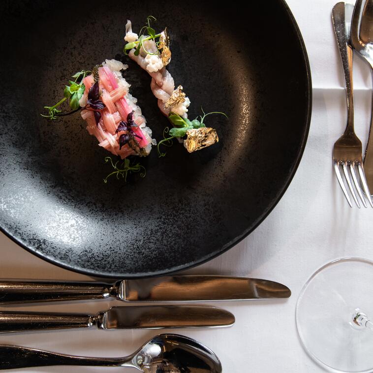 Gourmet-Vorspeise auf schwarzem Teller, dekoriert mit essbarem Blattgold, kunstvoll angerichtet und auf elegant gedecktem Tisch serviert