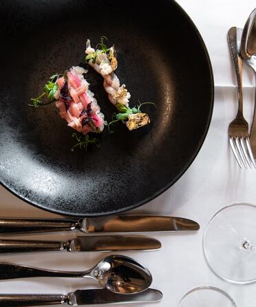Gourmet-Vorspeise auf schwarzem Teller, dekoriert mit essbarem Blattgold, kunstvoll angerichtet und auf elegant gedecktem Tisch serviert