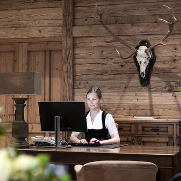 Rezeptionistin in traditioneller Tracht an einem Empfangstisch mit rustikaler Holzwand, Blumenschmuck und dekorativem Hirschgeweih | © SPORTALPEN