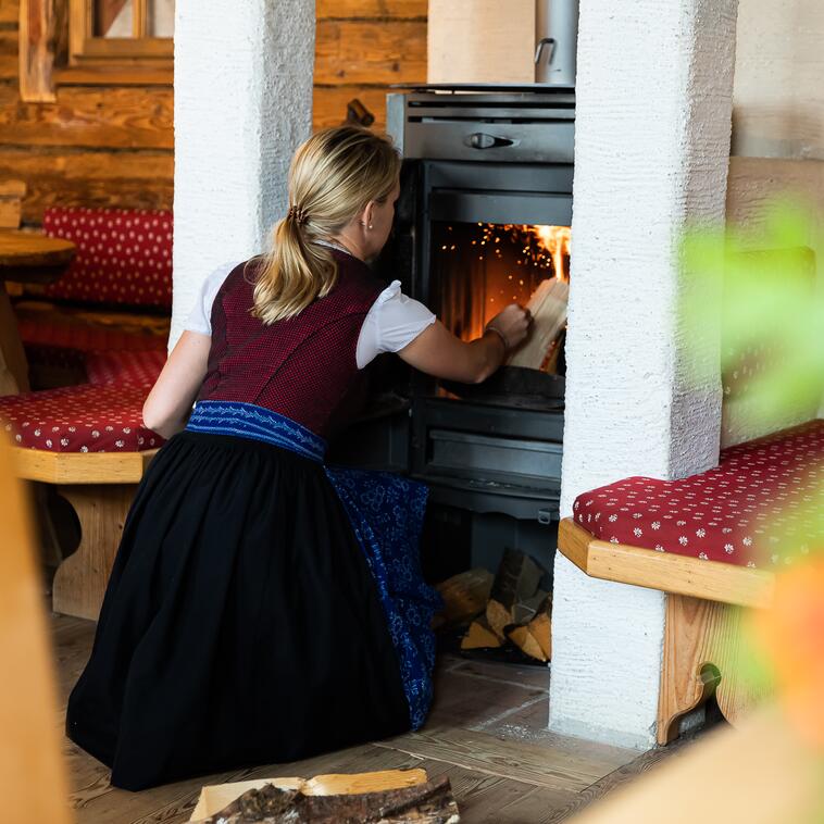 Eine Frau in traditioneller Tracht legt Holz in den Holzofen in einer Alm in Salzburg. 