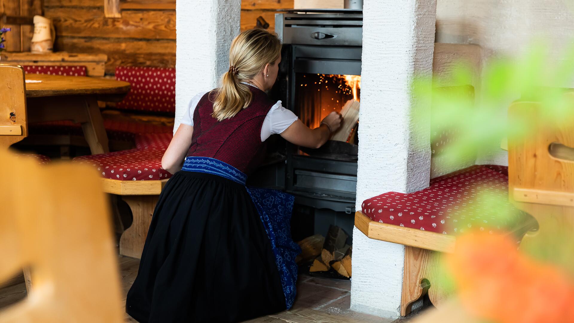 Eine Frau in traditioneller Tracht legt Holz in den Holzofen in einer Alm in Salzburg. 