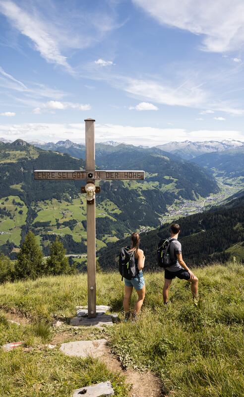 Zwei Wanderer vor Gipfelkreuz mit der Inschrift 'Ehre sei Gott in der Höhe' und Blick auf Berge und Täler | © www.fabianfranzferdinand.com